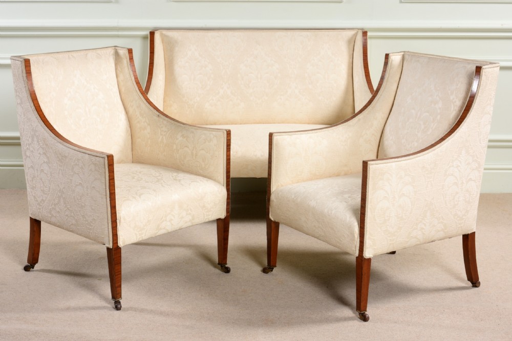 An antique three-piece Edwardian seating suite comprising a two-seater sofa and two matching armchairs upholstered in cream damask with mahogany frames and brass castors. View IV