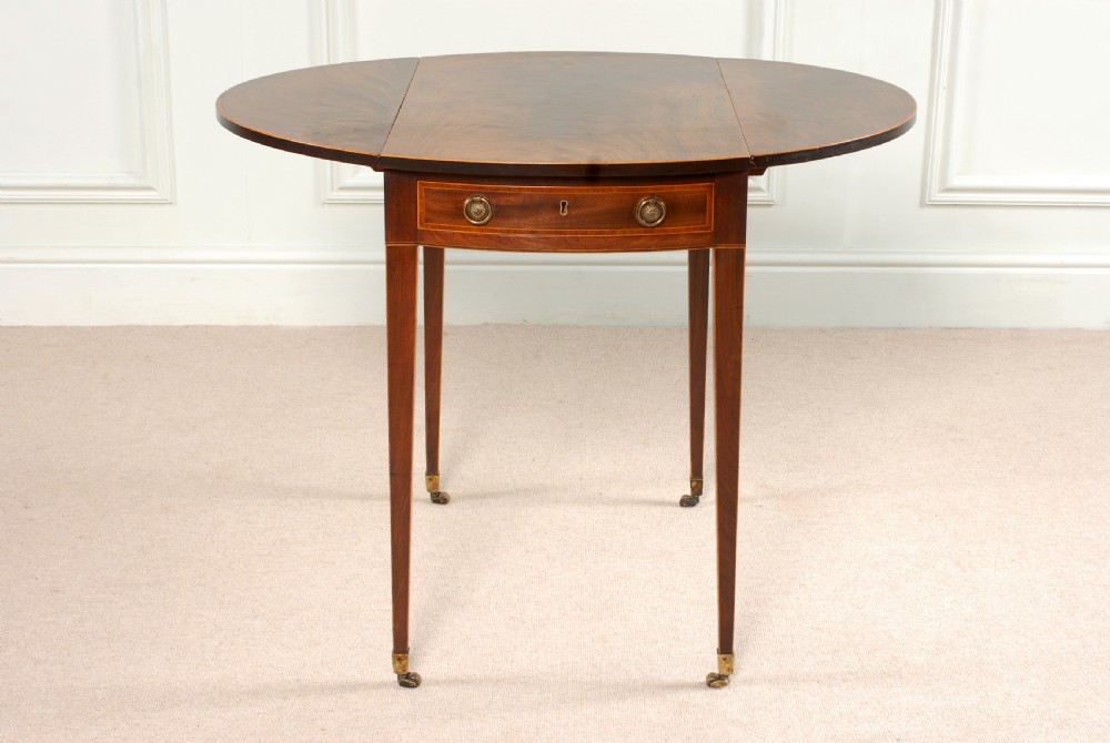 A Georgian Sheraton period mahogany oval Pembroke table with drop leaves, a single frieze drawer, boxwood inlay, and tapered legs ending in brass castors.