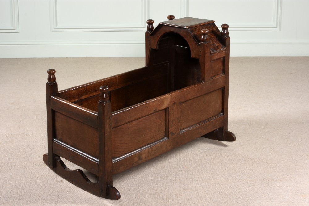 An Elizabethan dark oak rocking cradle with a panelled frame, hooded top section, turned finials, and curved wooden rockers.