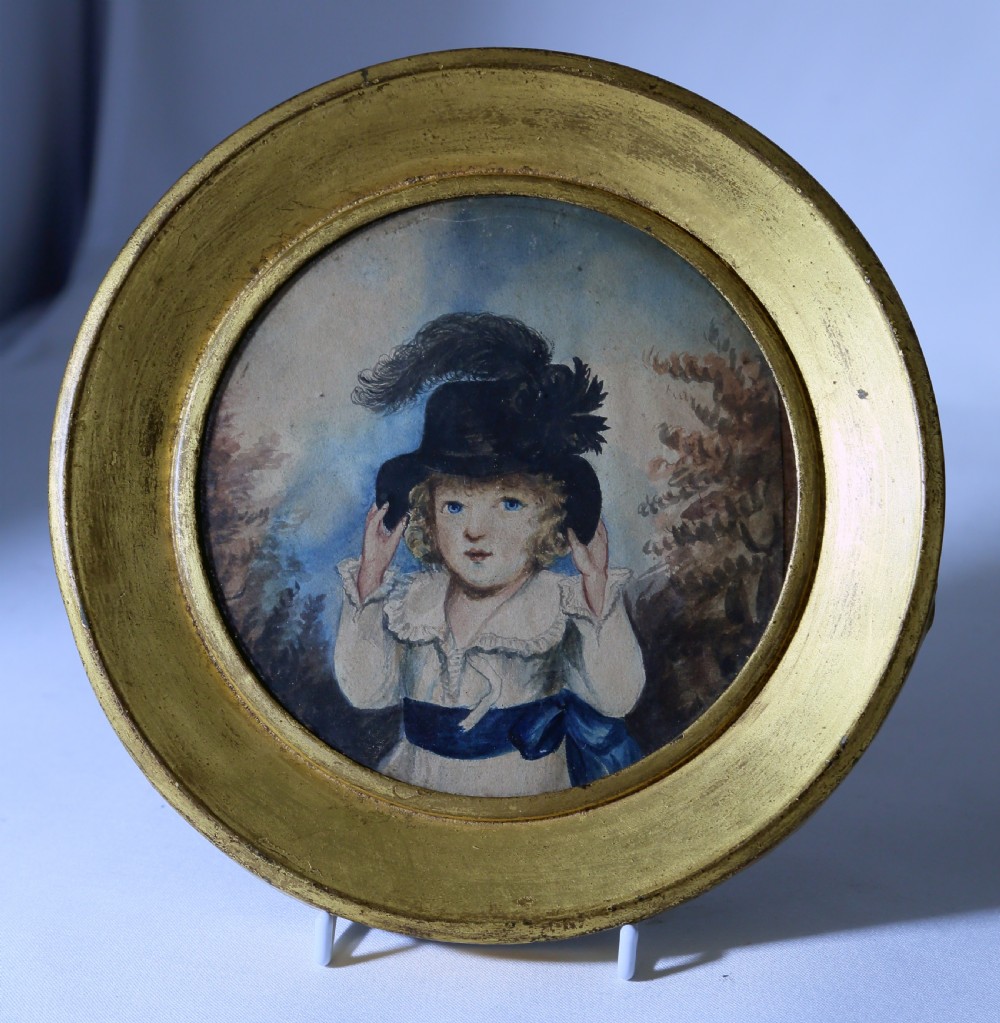 A circular watercolour portrait miniature of a young girl wearing a blue sash and black feathered hat, set within a round gilded wood frame.