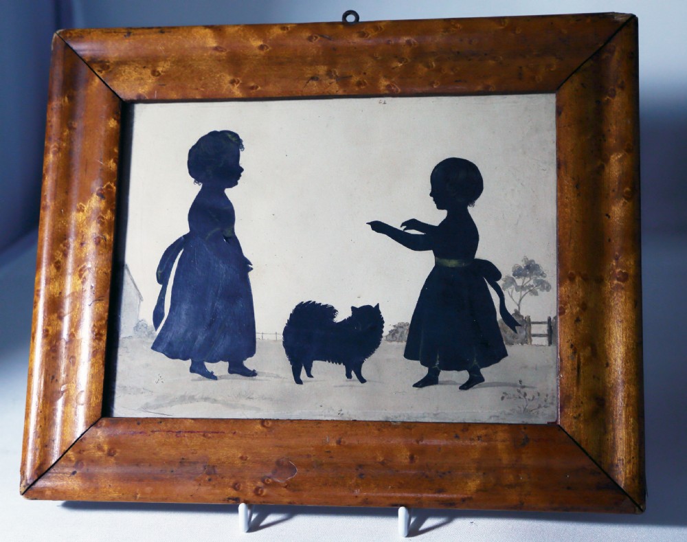 An antique silhouette portrait depicting two young children and a small dog in profile, set within a wide birds eye maple frame.