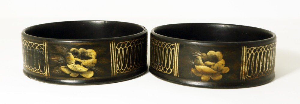 A pair of round black papier-mache coasters featuring hand-painted gold floral panels and looped decorative borders.