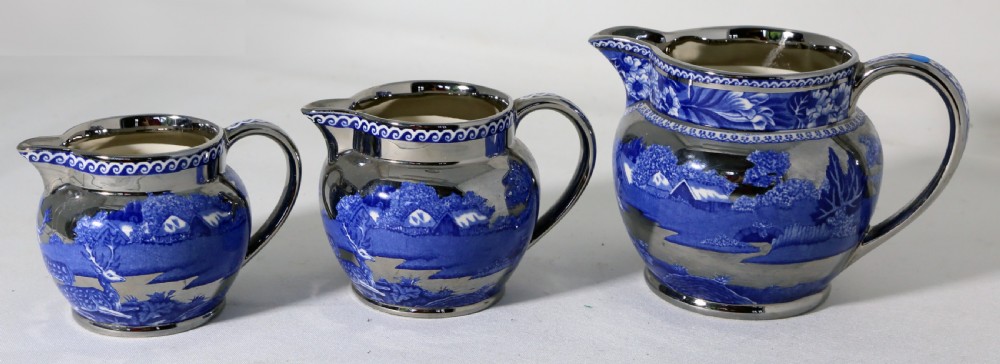 A set of three graduated blue earthenware jugs featuring a silver metallic lustre finish and a transfer-printed landscape scene with a fallow deer.