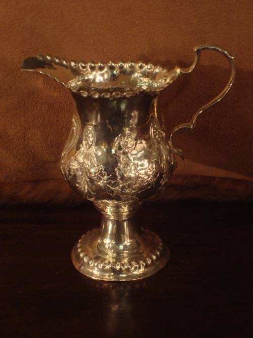 fig 1. A Georgian sterling silver pedestal cream jug featuring chased floral decoration, a scalloped rim, and a double-scroll handle.