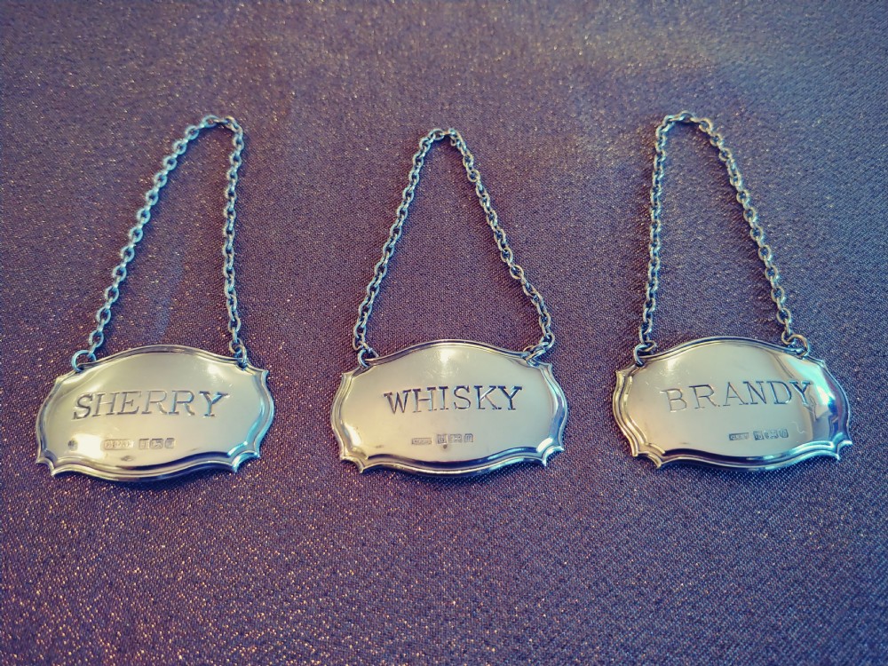 A set of three shaped sterling silver decanter labels engraved with Sherry, Whisky, and Brandy, suspended from fine link chains.