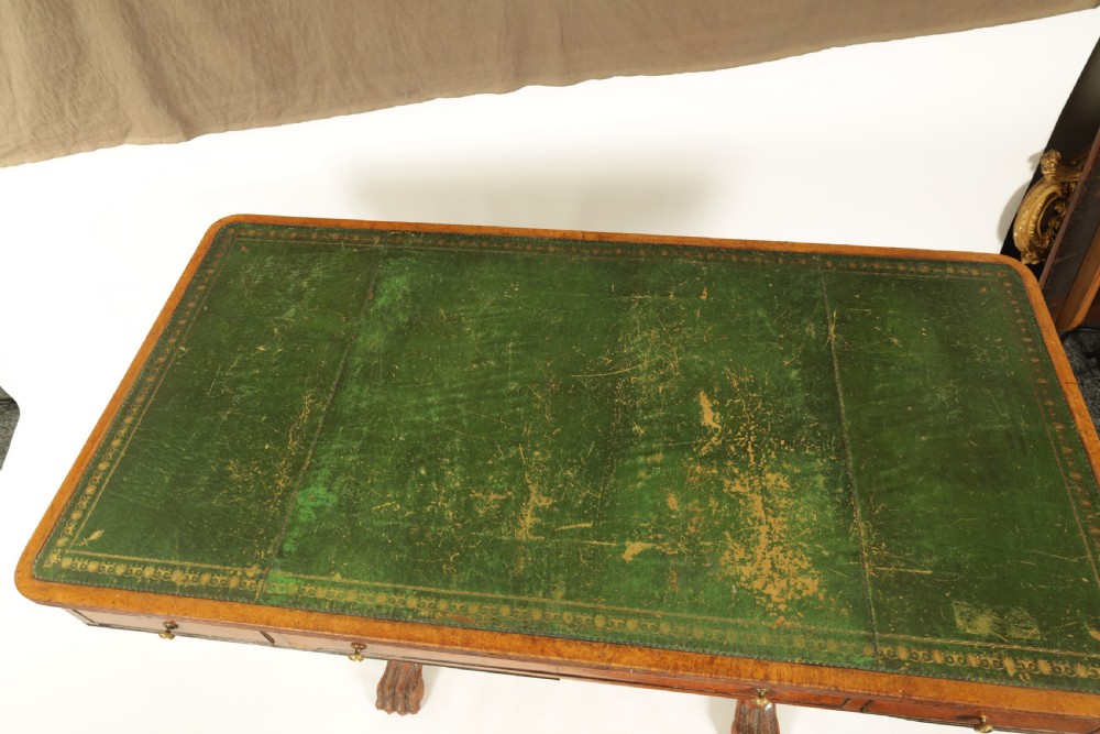 An antique amboyna and rosewood library table featuring a green leather inset top, frieze drawers with brass knobs, trestle legs, and carved lion paw feet. View VI