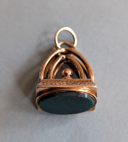 fig 1. An Edwardian 9ct gold watch fob featuring an arched cage superstructure, geometric chased detailing around the frame, and a dark green bloodstone seal base.