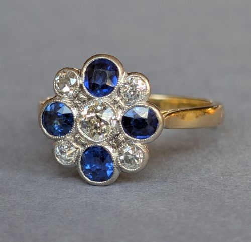 fig 1. An Art Deco cluster ring featuring a central round diamond surrounded by alternating blue sapphires and smaller diamonds in a flower formation set on a gold band.