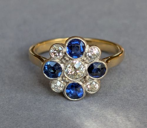 fig 2. An Art Deco cluster ring featuring a central round diamond surrounded by alternating blue sapphires and smaller diamonds in a flower formation set on a gold band.