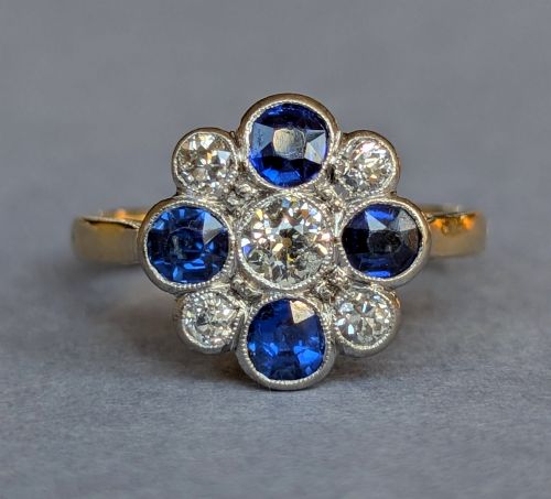 fig 3. An Art Deco cluster ring featuring a central round diamond surrounded by alternating blue sapphires and smaller diamonds in a flower formation set on a gold band.