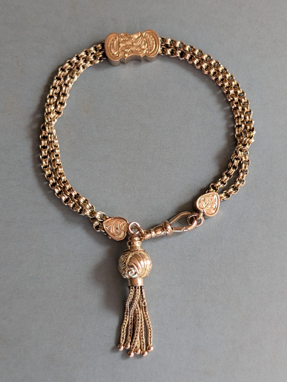 An antique 9ct gold double chain bracelet featuring engraved heart-shaped end pieces, a swivel clasp, and a hanging gold tassel charm.