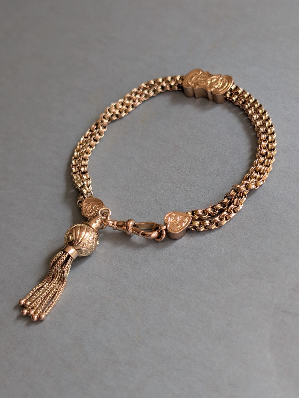 An antique 9ct gold double chain bracelet featuring engraved heart-shaped end pieces, a swivel clasp, and a hanging gold tassel charm. View VI