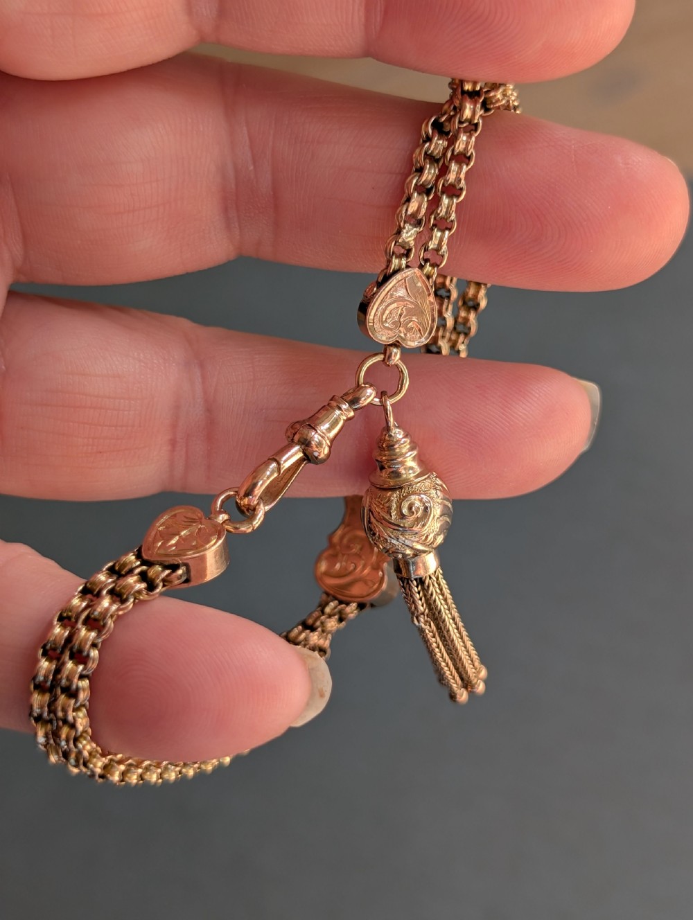 An antique 9ct gold double chain bracelet featuring engraved heart-shaped end pieces, a swivel clasp, and a hanging gold tassel charm. View X