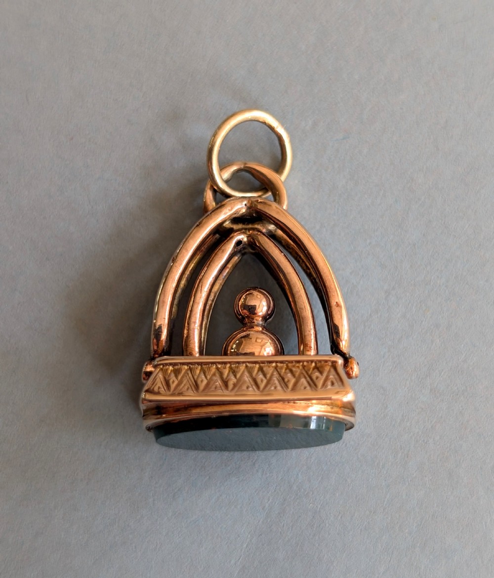 An Edwardian 9ct gold watch fob featuring an arched cage superstructure, geometric chased detailing around the frame, and a dark green bloodstone seal base.