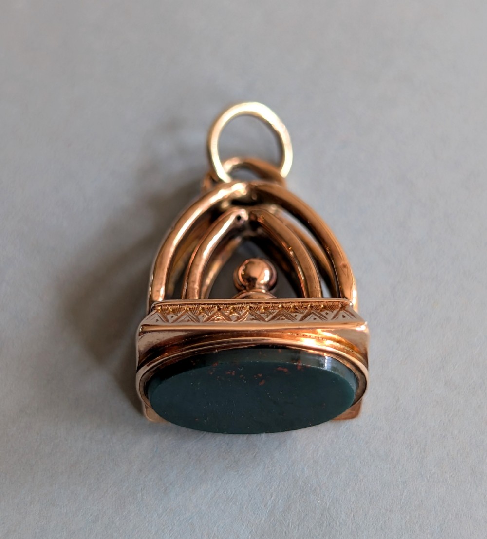 An Edwardian 9ct gold watch fob featuring an arched cage superstructure, geometric chased detailing around the frame, and a dark green bloodstone seal base. View II