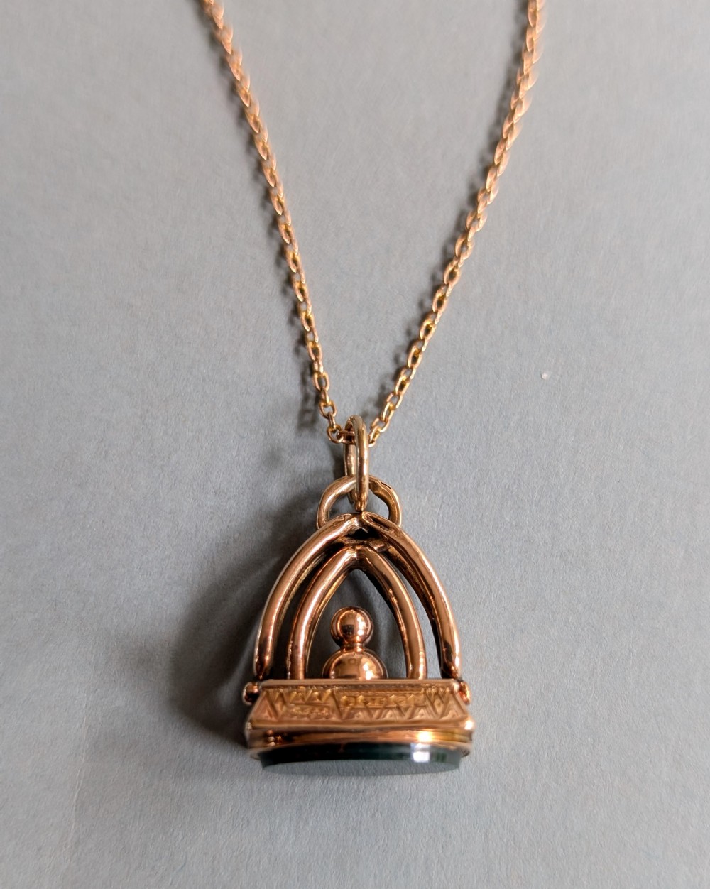 An Edwardian 9ct gold watch fob featuring an arched cage superstructure, geometric chased detailing around the frame, and a dark green bloodstone seal base. View VII
