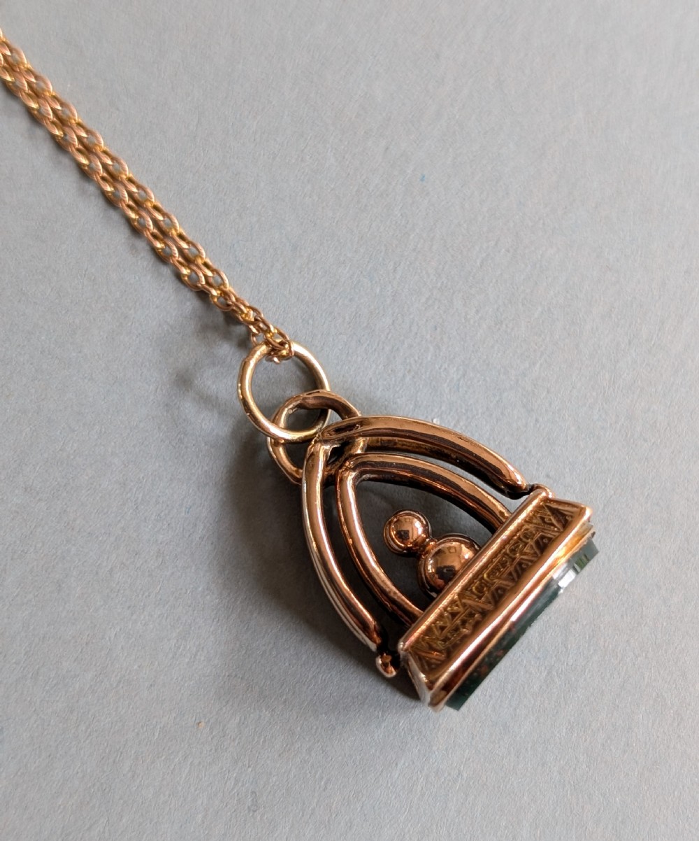 An Edwardian 9ct gold watch fob featuring an arched cage superstructure, geometric chased detailing around the frame, and a dark green bloodstone seal base. View X