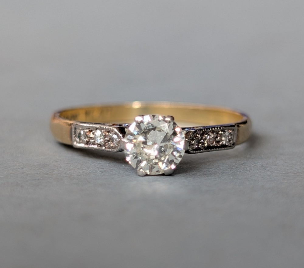 An Art Deco diamond solitaire engagement ring featuring a central round diamond set in platinum claws with diamond-accented shoulders on a yellow gold band.