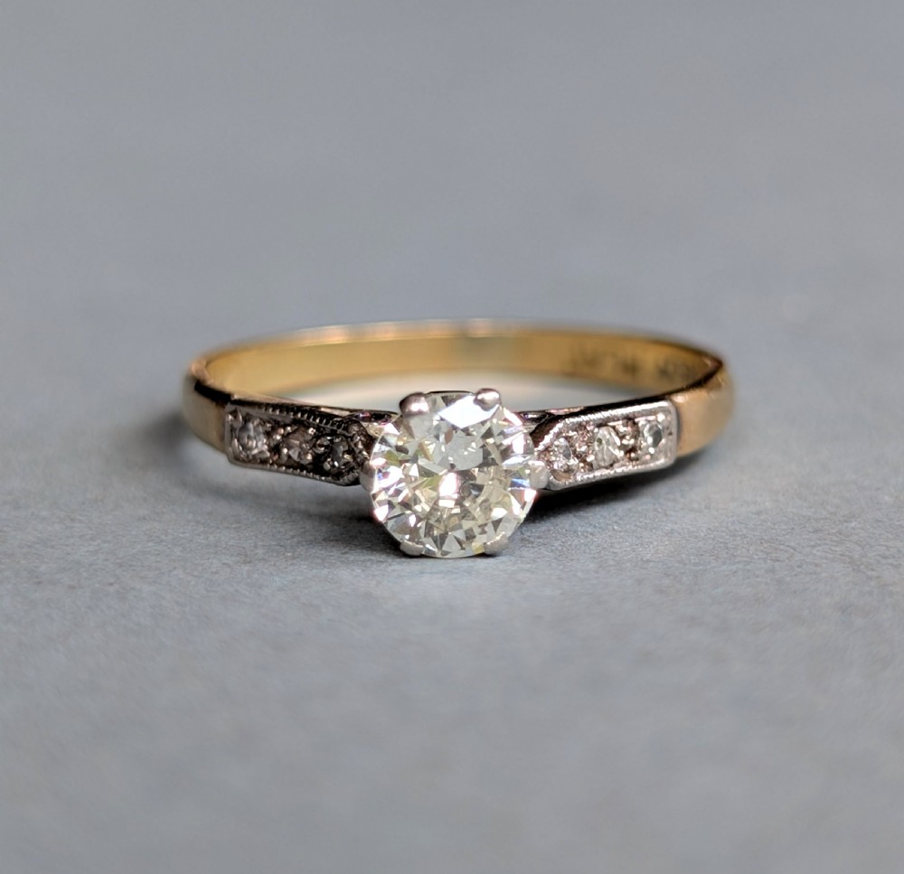 An Art Deco diamond solitaire engagement ring featuring a central round diamond set in platinum claws with diamond-accented shoulders on a yellow gold band. View VI