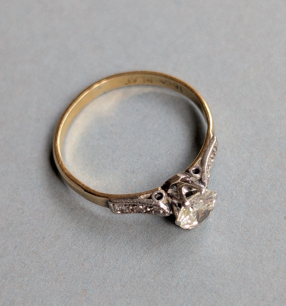 An Art Deco diamond solitaire engagement ring featuring a central round diamond set in platinum claws with diamond-accented shoulders on a yellow gold band. View X