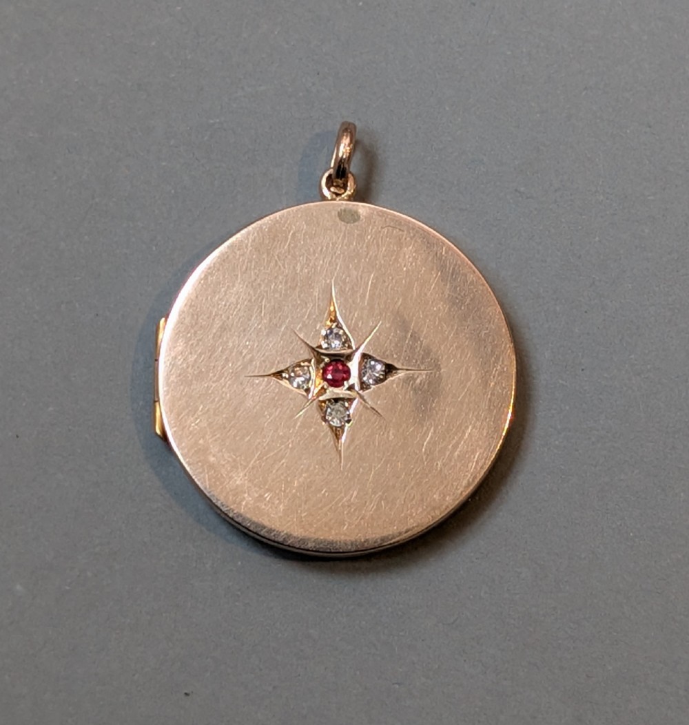 A circular 9ct gold locket featuring a starburst engraving set with a central red stone and surrounding clear paste stones.