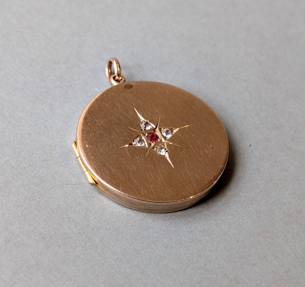 A circular 9ct gold locket featuring a starburst engraving set with a central red stone and surrounding clear paste stones. View II