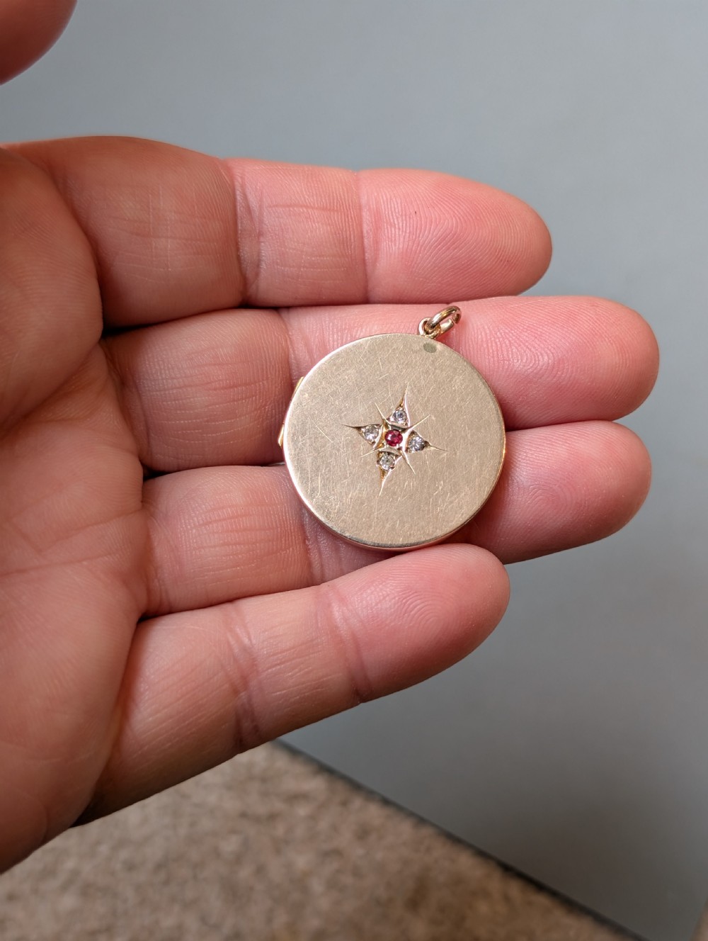 A circular 9ct gold locket featuring a starburst engraving set with a central red stone and surrounding clear paste stones. View V