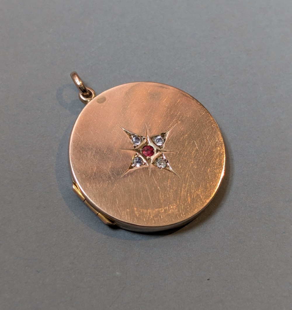 A circular 9ct gold locket featuring a starburst engraving set with a central red stone and surrounding clear paste stones. View IX