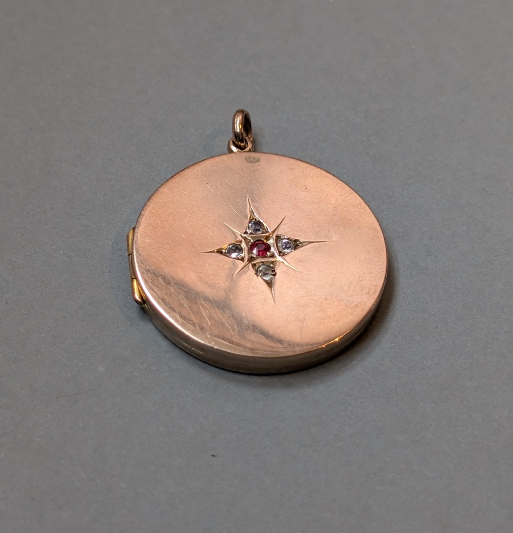 A circular 9ct gold locket featuring a starburst engraving set with a central red stone and surrounding clear paste stones. View X