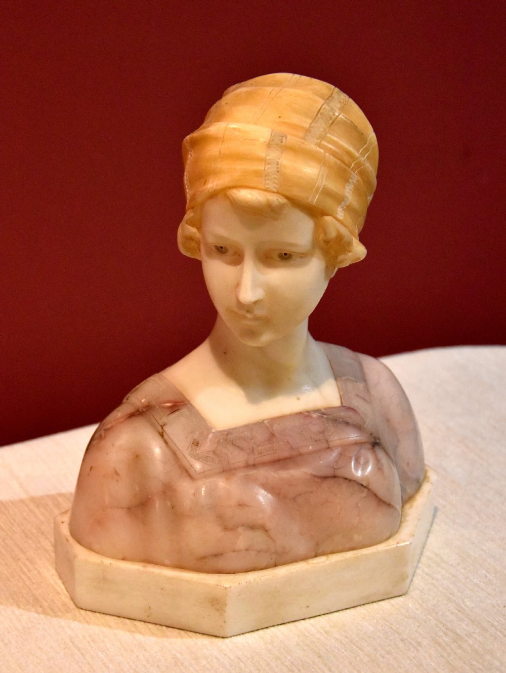 A carved alabaster and pink marble bust of a young woman wearing a draped cap set upon an octagonal white base.