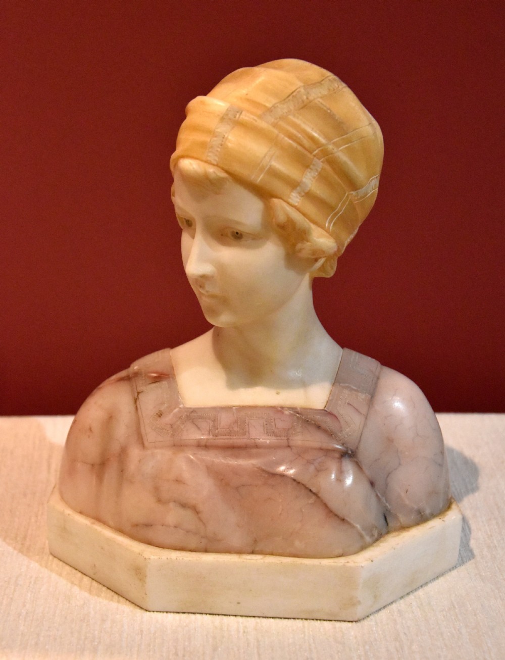 A carved alabaster and pink marble bust of a young woman wearing a draped cap set upon an octagonal white base. View II