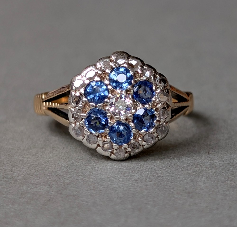 An Art Deco 18ct yellow gold ring featuring a central diamond encircled by six blue sapphires and an outer halo of small diamonds.