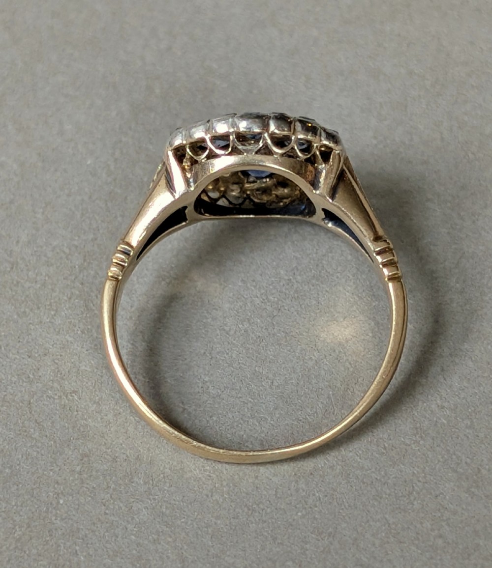 An Art Deco 18ct yellow gold ring featuring a central diamond encircled by six blue sapphires and an outer halo of small diamonds. View VI