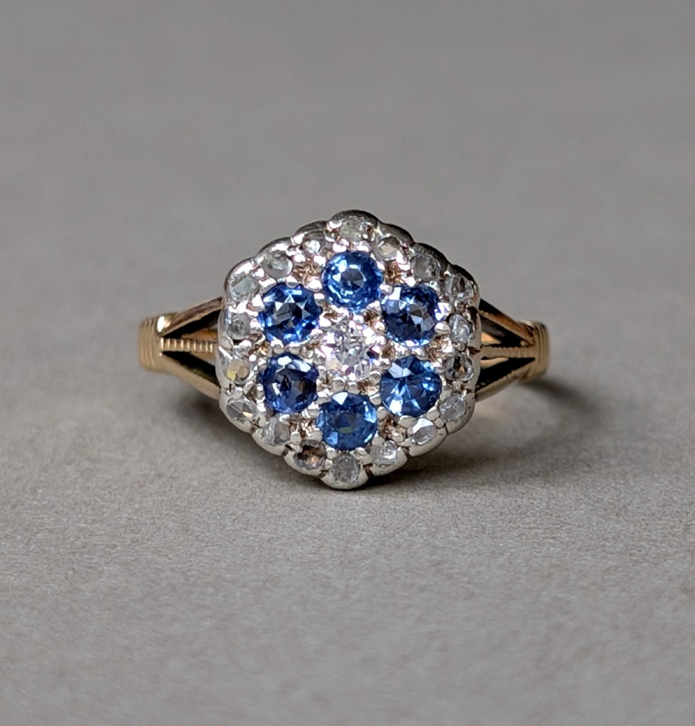 An Art Deco 18ct yellow gold ring featuring a central diamond encircled by six blue sapphires and an outer halo of small diamonds. View VIII