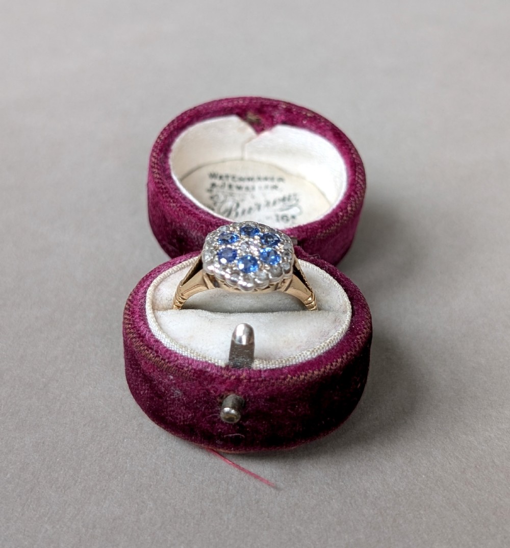 An Art Deco 18ct yellow gold ring featuring a central diamond encircled by six blue sapphires and an outer halo of small diamonds. View X