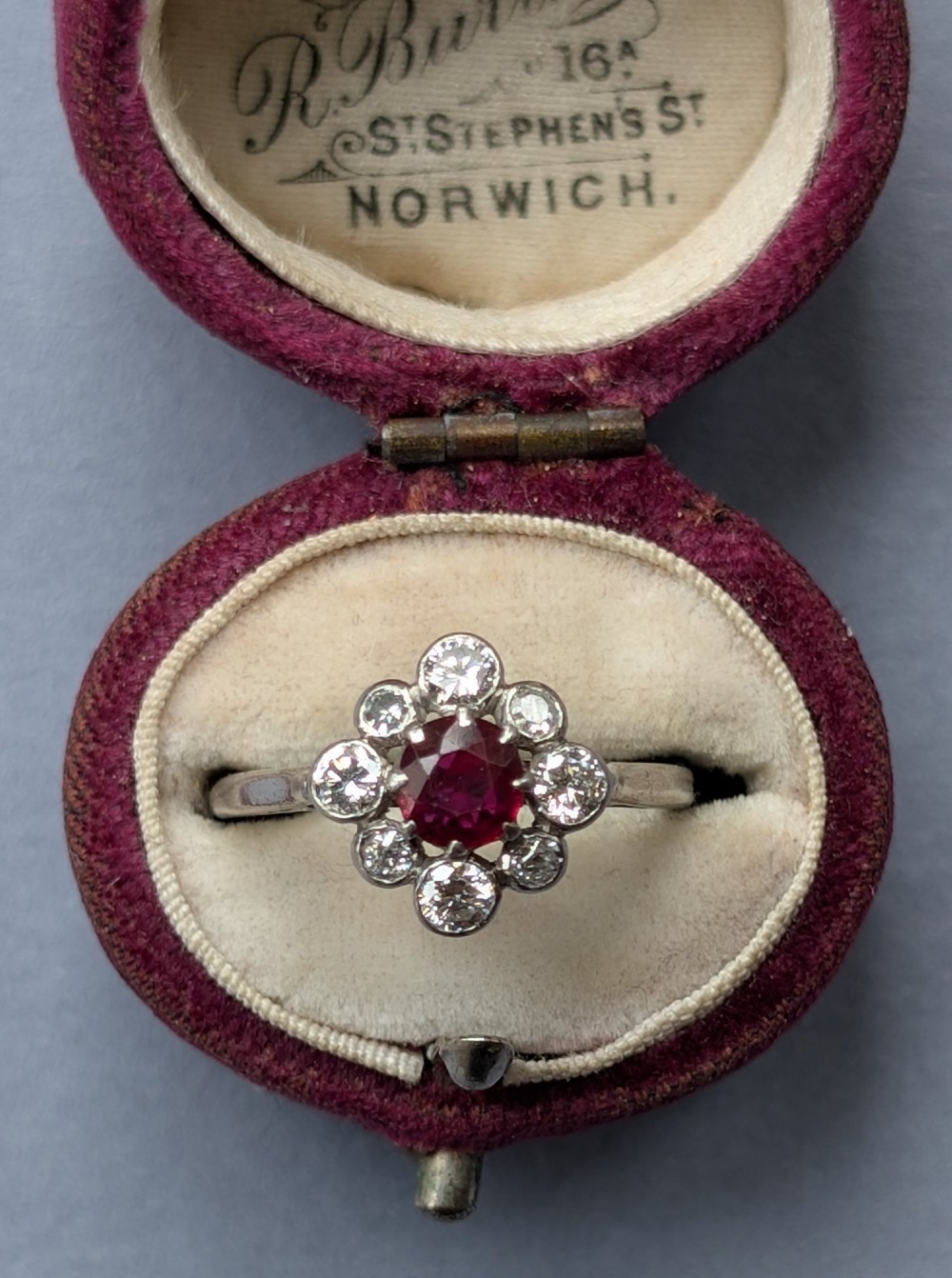 An Art Deco platinum ring featuring a circular central red ruby surrounded by a cluster halo of eight round diamonds. View VI
