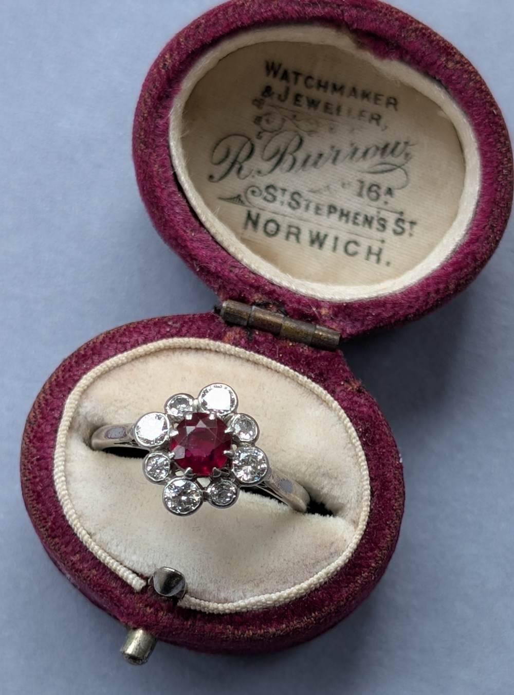 An Art Deco platinum ring featuring a circular central red ruby surrounded by a cluster halo of eight round diamonds. View VII