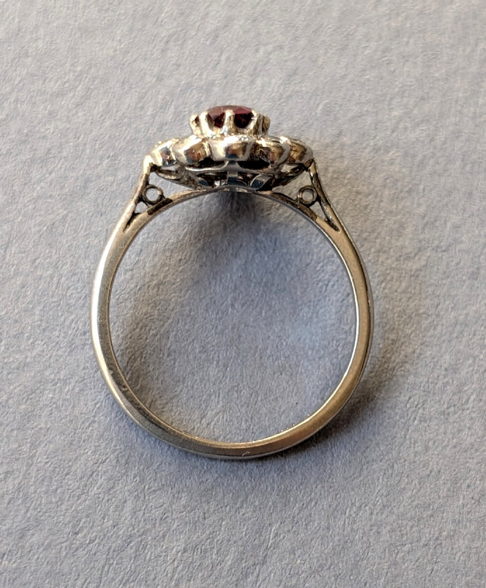 An Art Deco platinum ring featuring a circular central red ruby surrounded by a cluster halo of eight round diamonds. View X