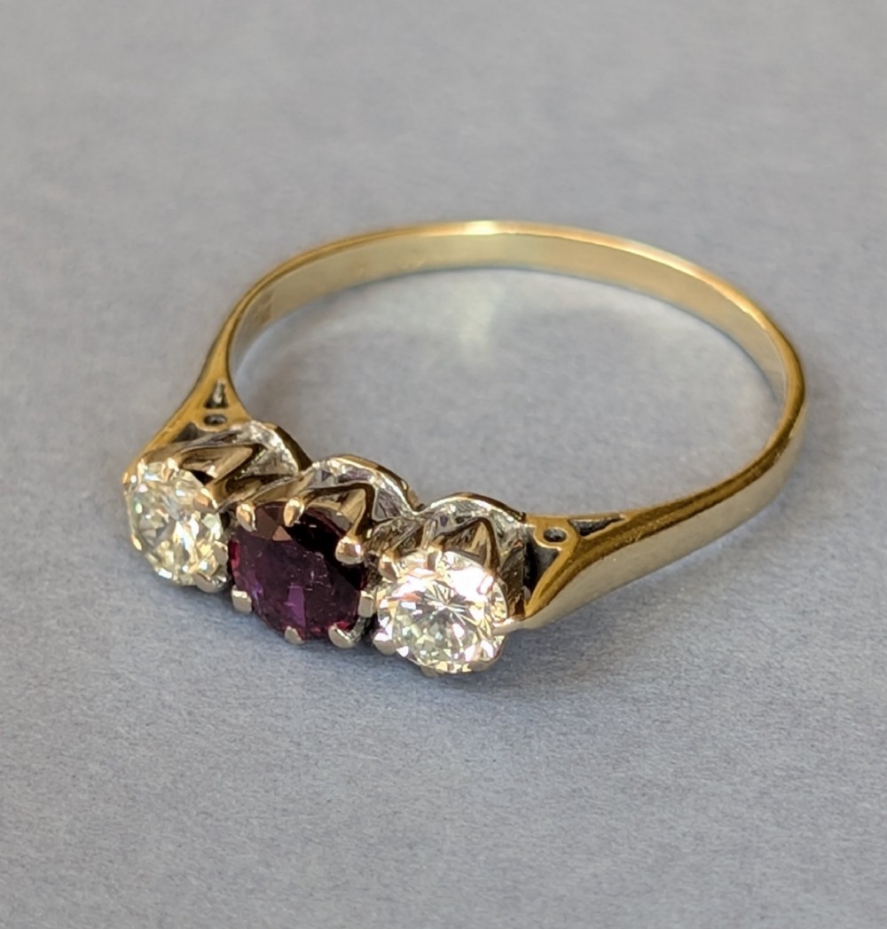 An 18ct gold trilogy ring featuring a round central red ruby flanked by two diamonds in claw settings. View VI