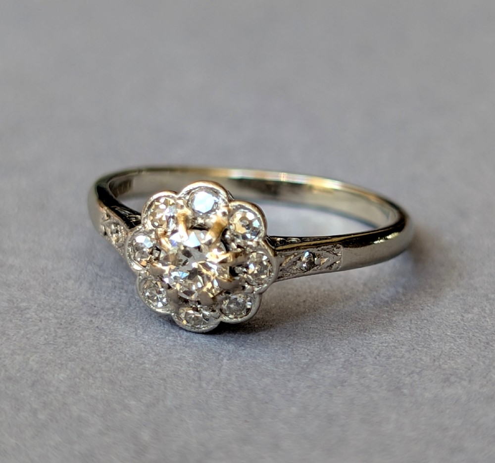 An Art Deco diamond daisy cluster ring with a circular central diamond surrounded by eight smaller diamonds set on a polished band with detailed shoulders. View III