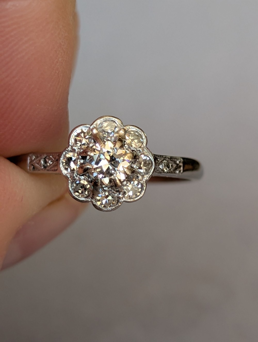 An Art Deco diamond daisy cluster ring with a circular central diamond surrounded by eight smaller diamonds set on a polished band with detailed shoulders. View IV