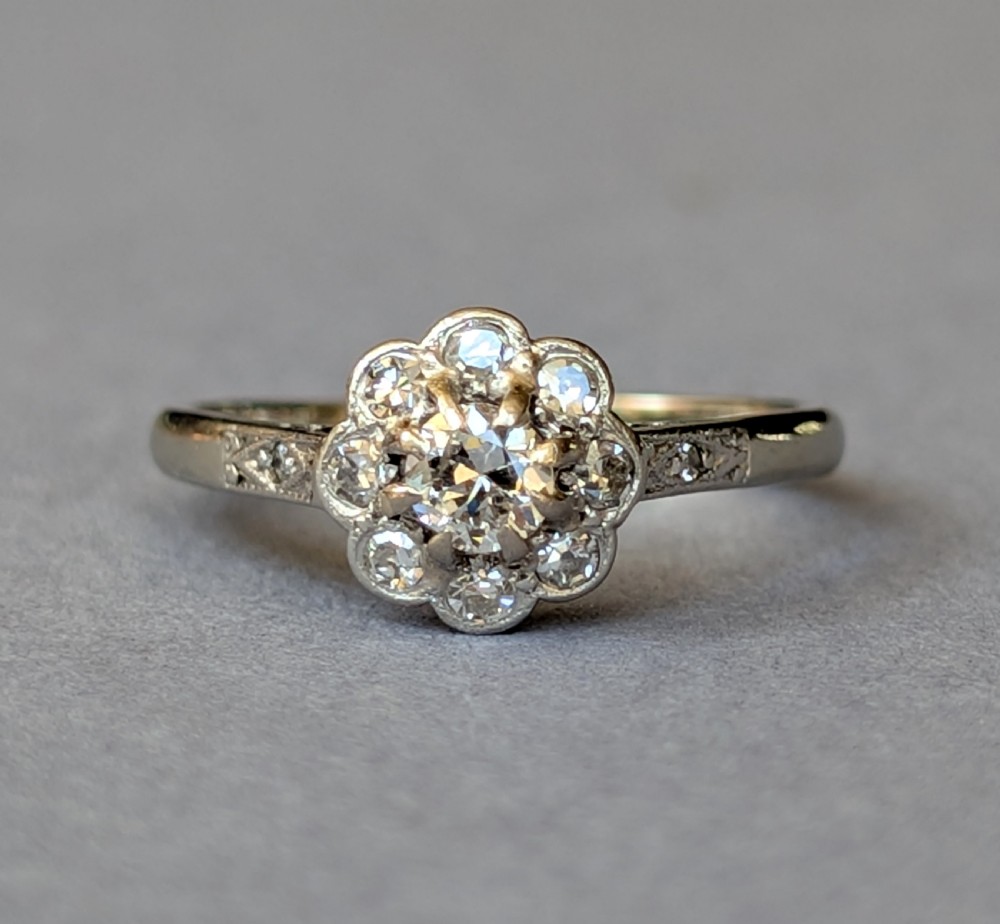 An Art Deco diamond daisy cluster ring with a circular central diamond surrounded by eight smaller diamonds set on a polished band with detailed shoulders. View V