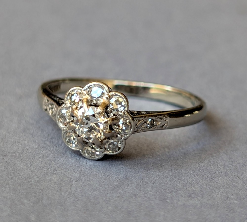 An Art Deco diamond daisy cluster ring with a circular central diamond surrounded by eight smaller diamonds set on a polished band with detailed shoulders. View VI