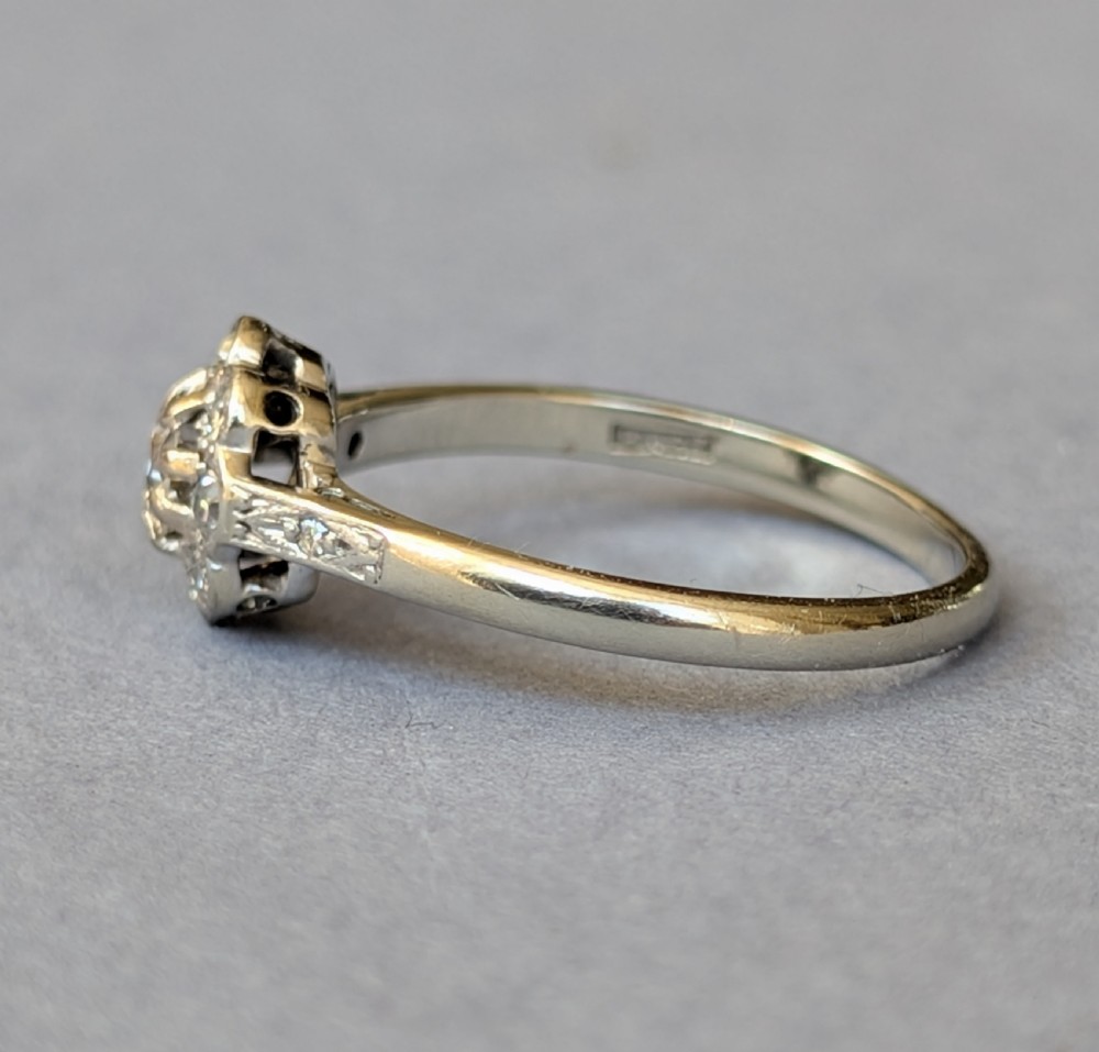 An Art Deco diamond daisy cluster ring with a circular central diamond surrounded by eight smaller diamonds set on a polished band with detailed shoulders. View IX
