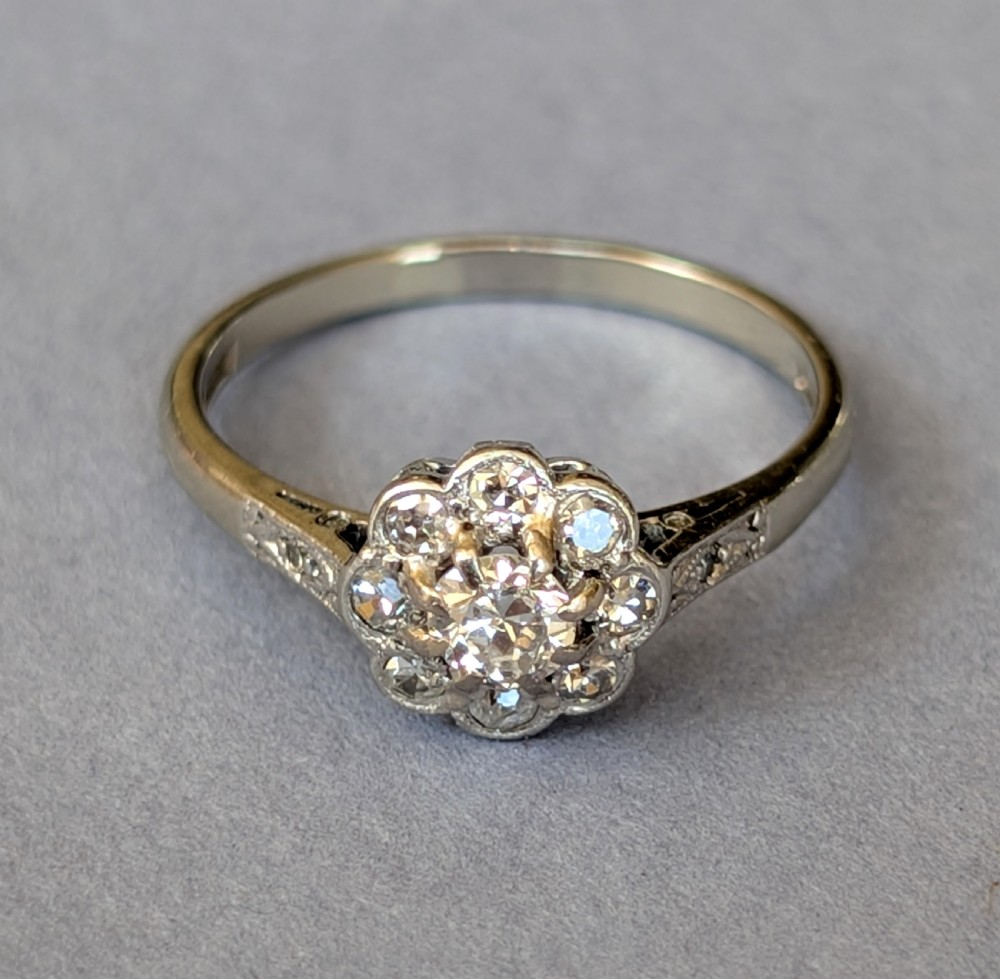 An Art Deco diamond daisy cluster ring with a circular central diamond surrounded by eight smaller diamonds set on a polished band with detailed shoulders. View X