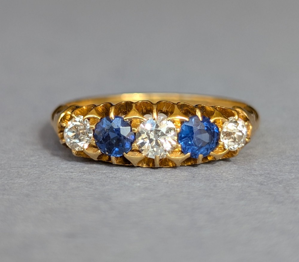 An Edwardian 18ct yellow gold five-stone ring featuring alternating blue sapphires and old cut diamonds set within a carved scroll head.