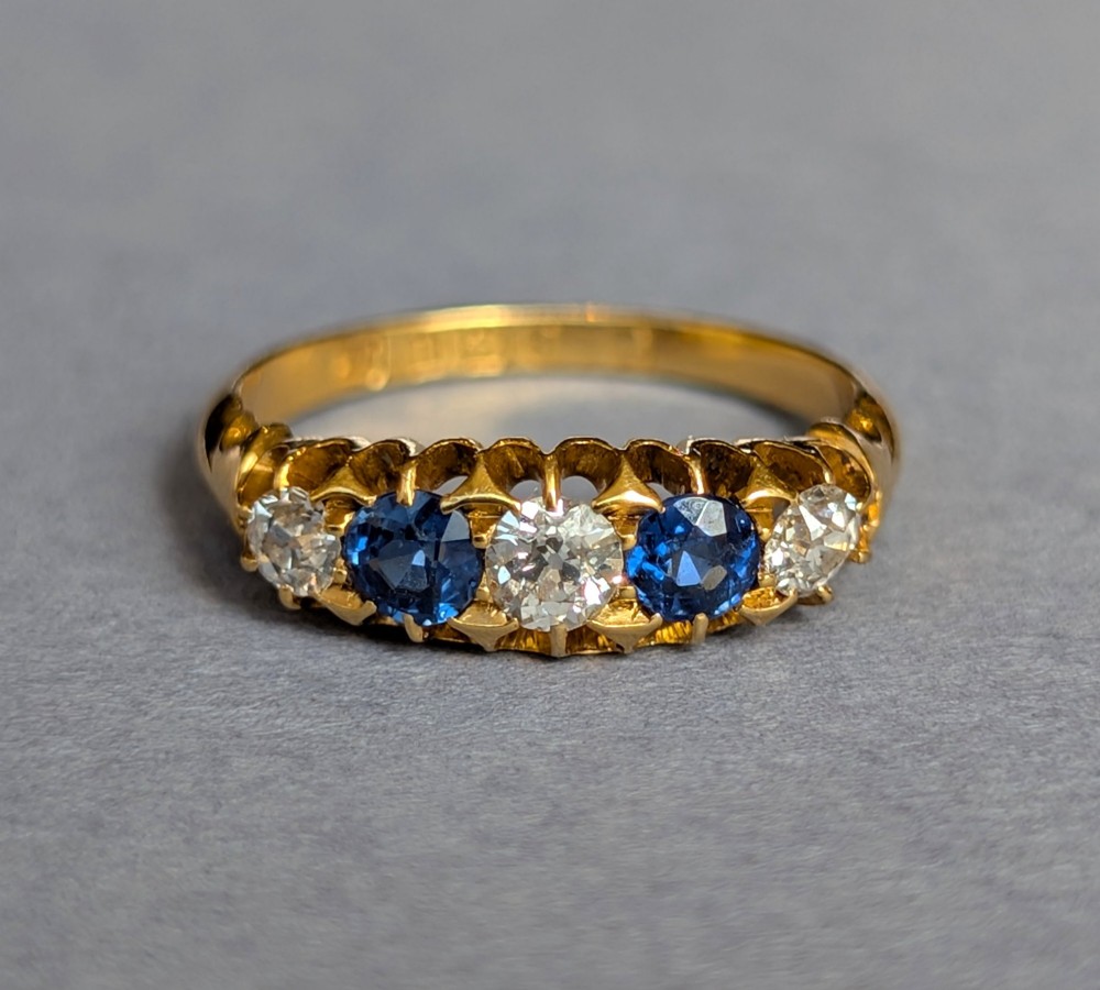 An Edwardian 18ct yellow gold five-stone ring featuring alternating blue sapphires and old cut diamonds set within a carved scroll head. View II