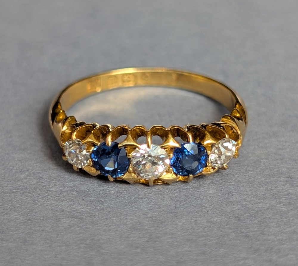An Edwardian 18ct yellow gold five-stone ring featuring alternating blue sapphires and old cut diamonds set within a carved scroll head. View IV