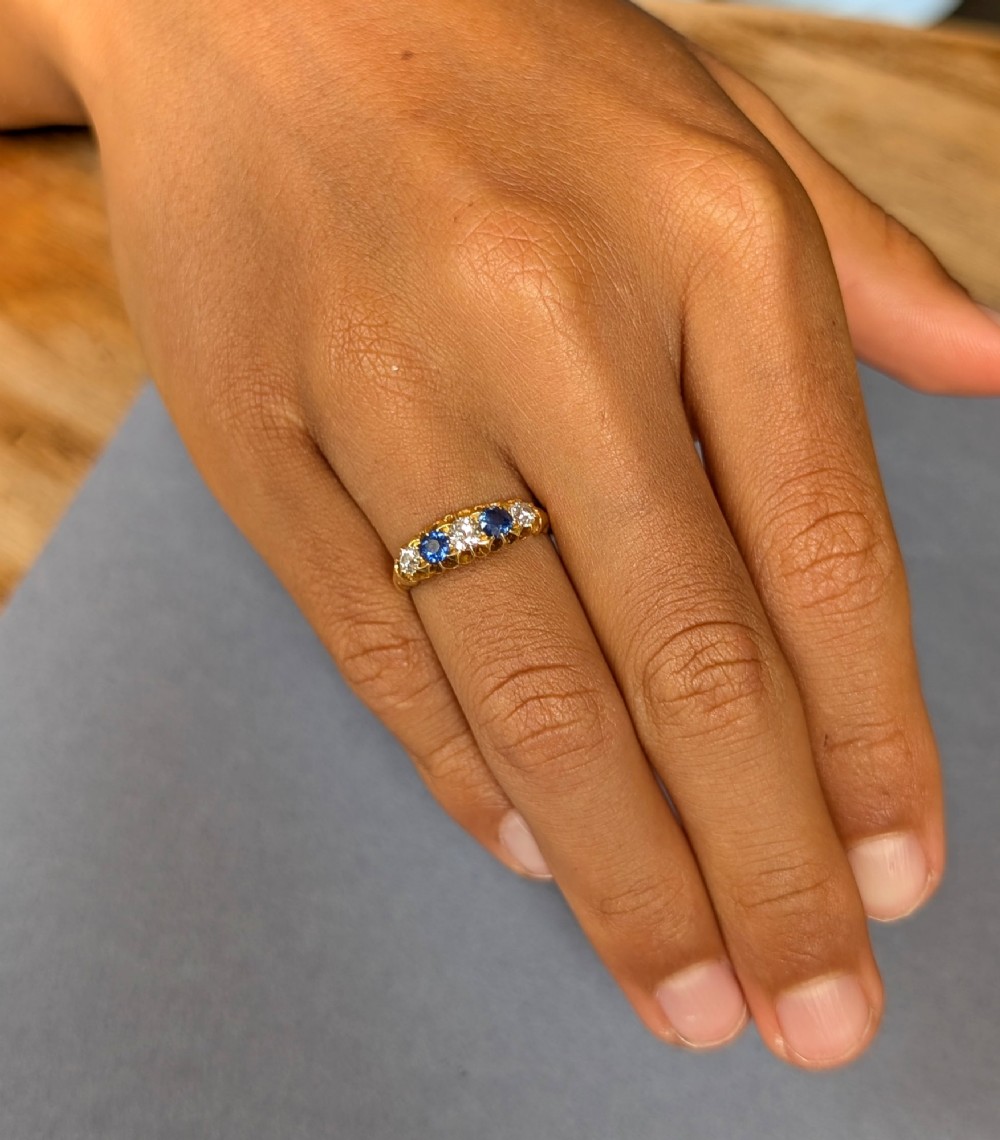 An Edwardian 18ct yellow gold five-stone ring featuring alternating blue sapphires and old cut diamonds set within a carved scroll head. View V