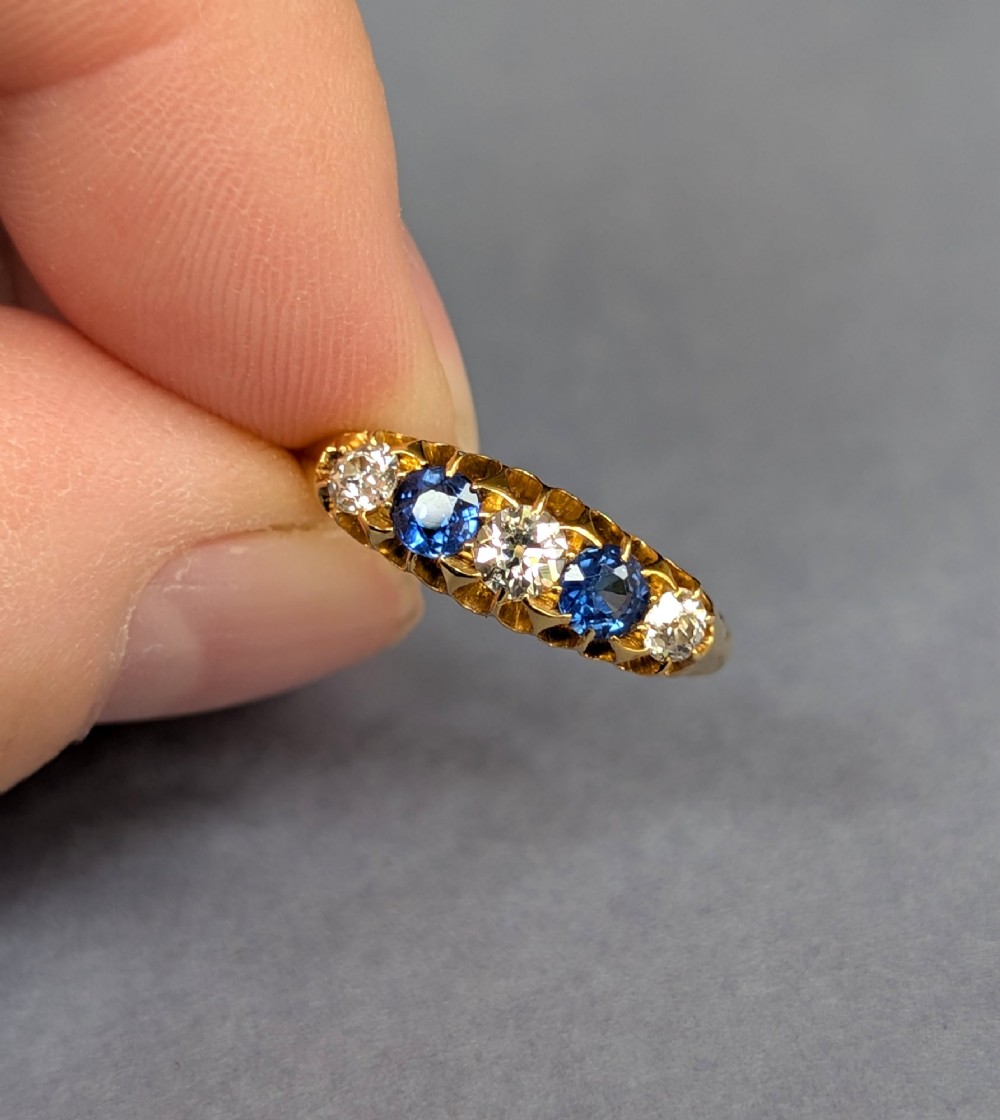 An Edwardian 18ct yellow gold five-stone ring featuring alternating blue sapphires and old cut diamonds set within a carved scroll head. View VII